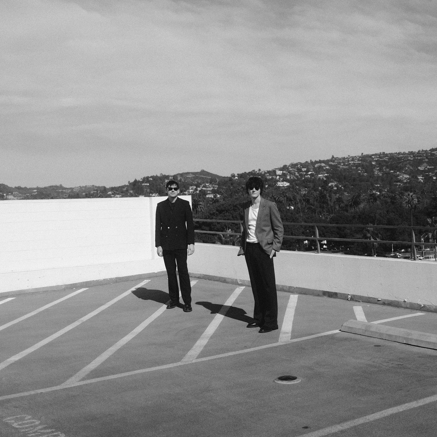 Black and white photo of the duo in sunglasses on a rooftop car park above the hills
